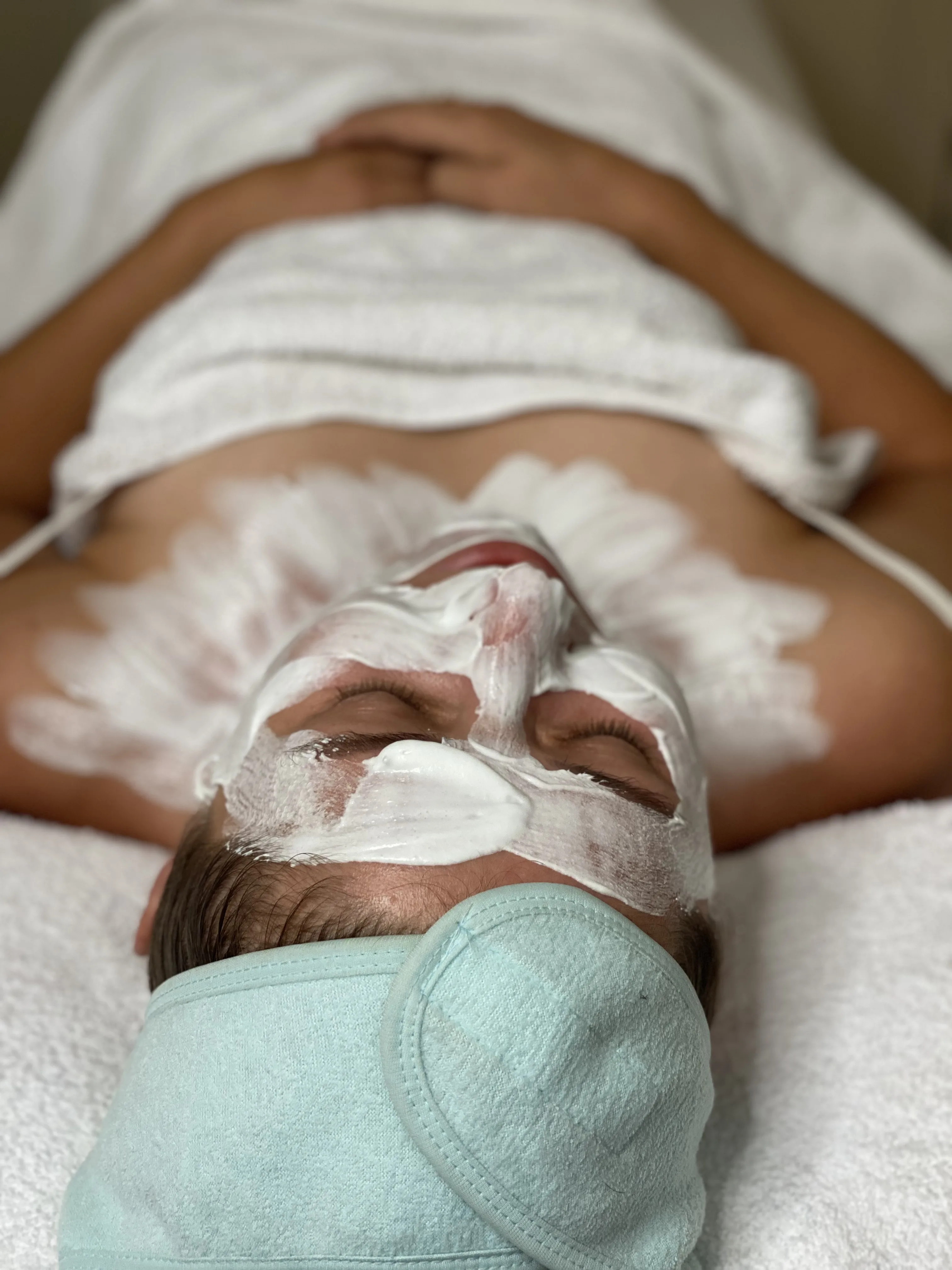 Client with full white facial mask and mint eye pads on the spa bed — facial treatment in progress